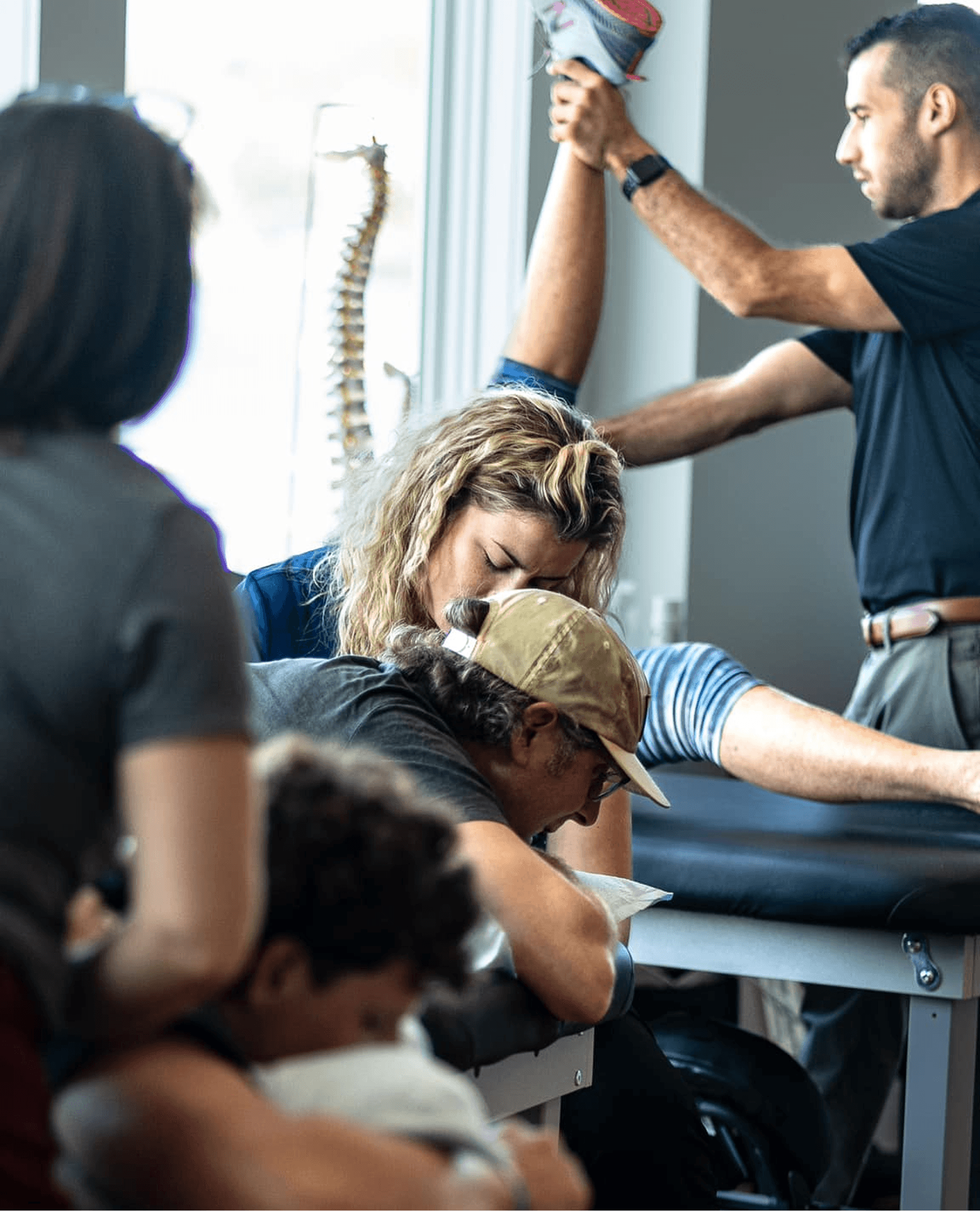Busy rehab setting where a female therapist supports a patient prone on a treatment table while another therapist in the background lifts and stretches a client’s leg.