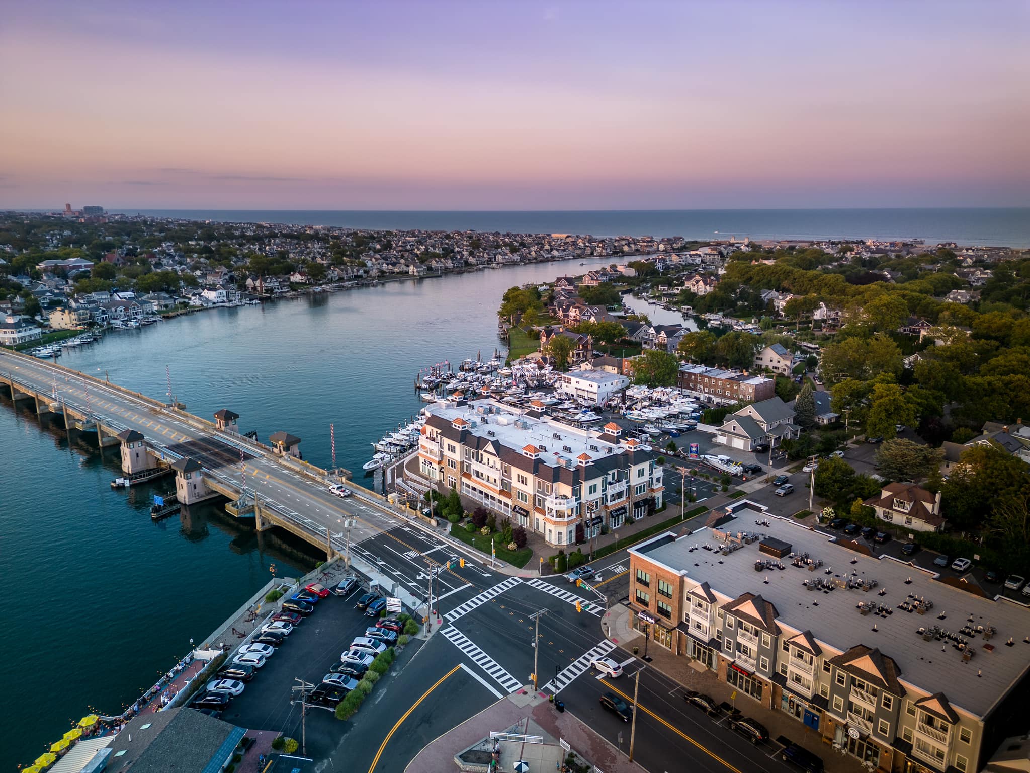 Aerial view of a bridge, a marina and a shore town at dusk, with the ocean beyond
